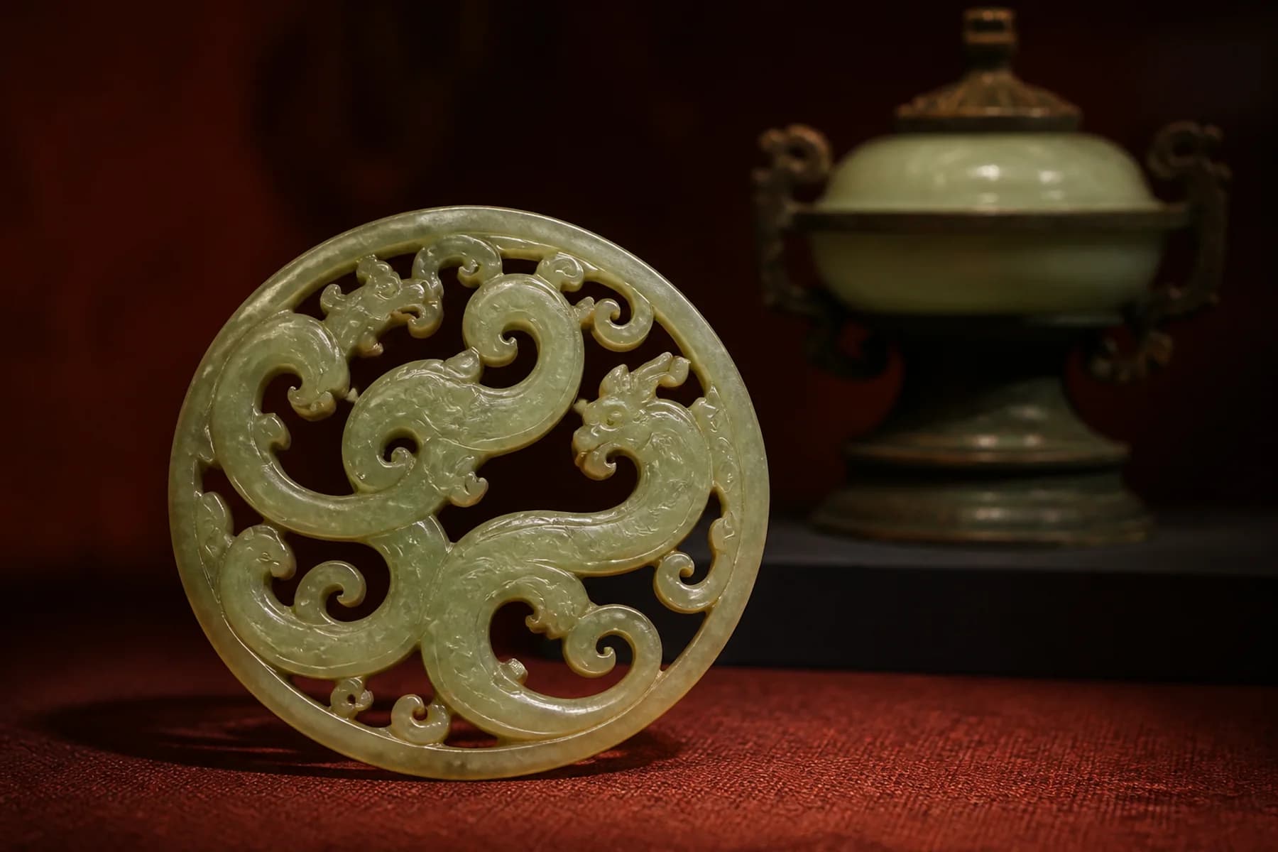 Bodegón editorial de museo con un colgante calado de jade verde pálido y una vasija ritual desenfocada al fondo