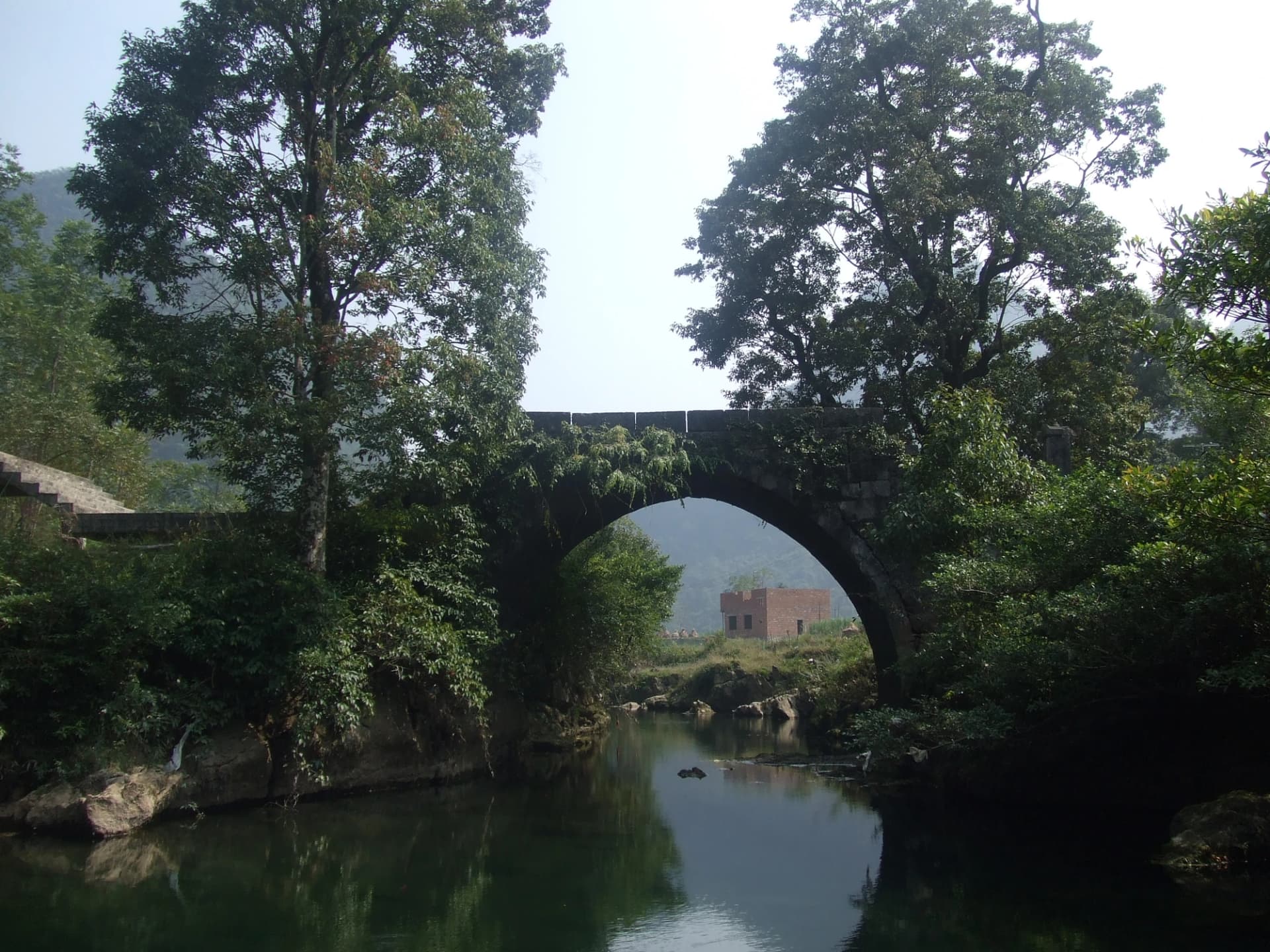 Un antiguo puente de piedra entre árboles y una aldea de Maolan